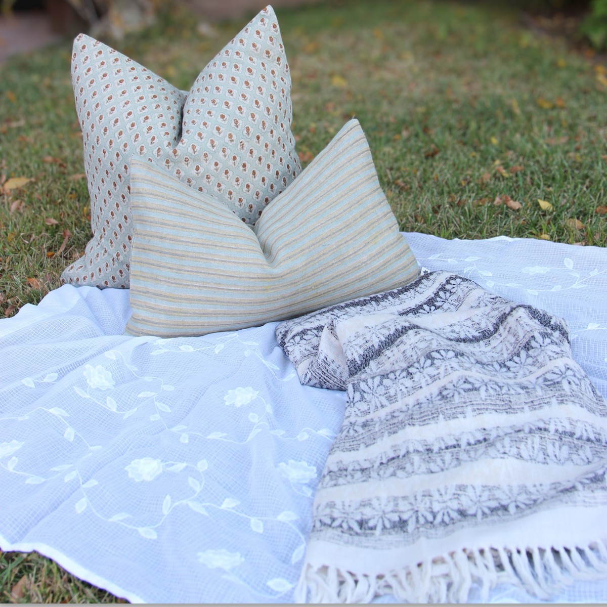 Handwoven gray and white floral throw with fringe, styled with a sage hand block printed cushion and a striped cushion on grass